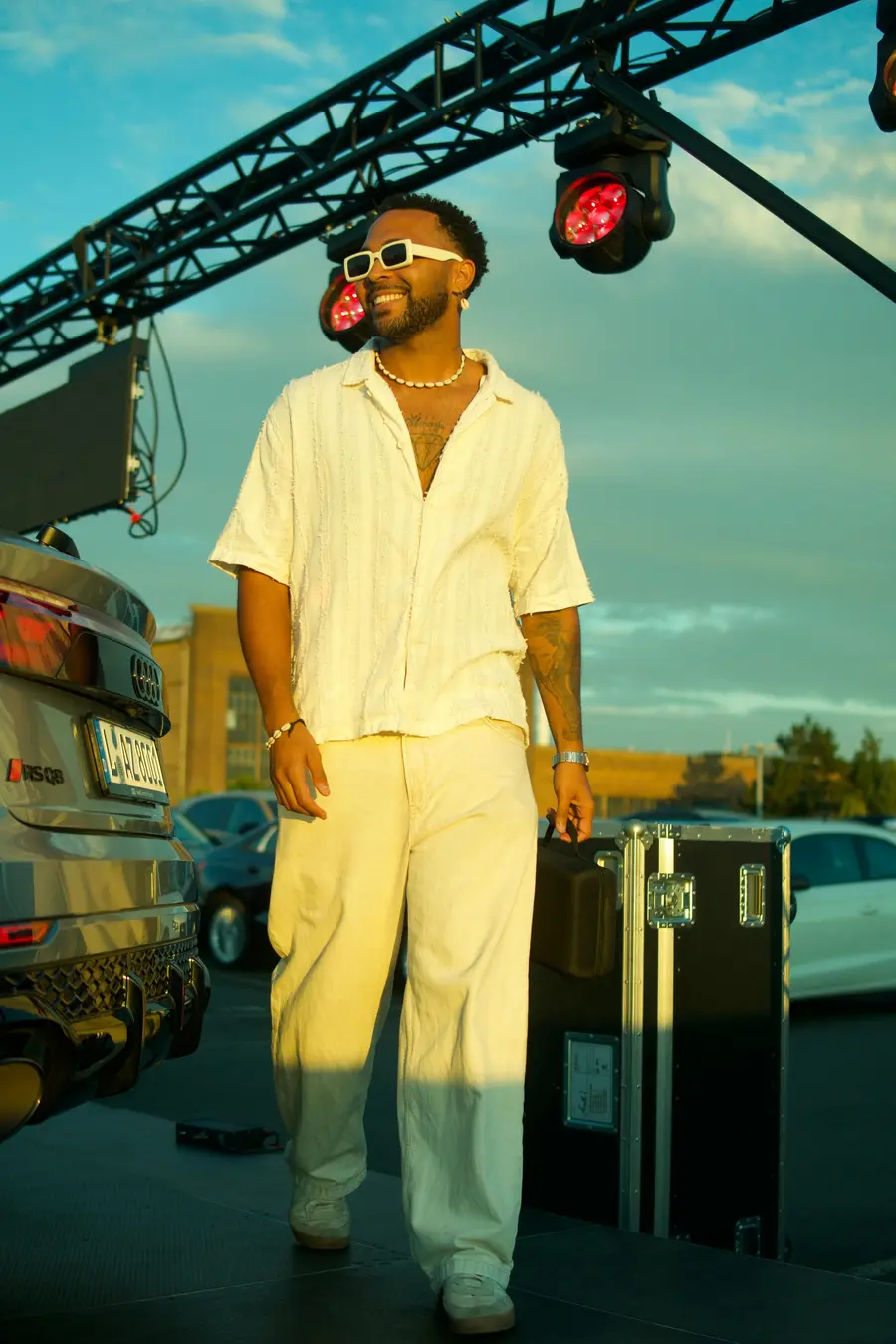 SEKOU in cream linen beside a stage truss at sunset